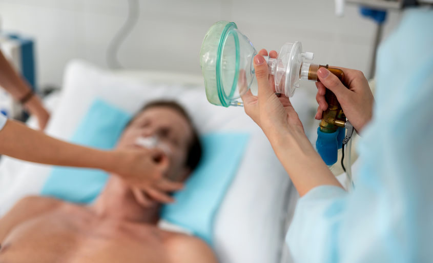 Close up of female hands holding medical respiratory equipment. Man lying on hospital bed and doctor arm on blurred background